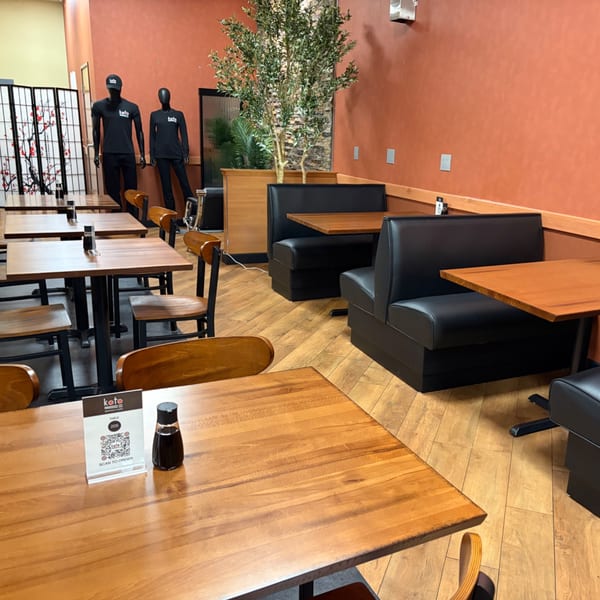 Warmly lit restaurant dining area with wooden tables, black booths, decorative plants, and branded mannequins near a folding screen.