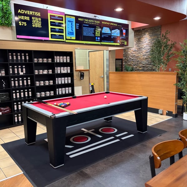 Restaurant interior featuring a red-felt pool table, merchandise shelves, digital menu screens, wood paneling, and decorative plants.